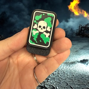 May include: A black and green patch with a white skull and crossbones design. The patch is held in a hand against a background of a cracked, dry surface.