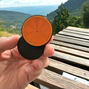 May include: A black and orange round phone grip with a geometric pattern. The grip is designed to be attached to the back of a phone.