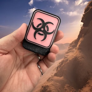 May include: A black and pink biohazard symbol patch with a black border. The patch is held in a hand against a background of sand dunes.