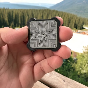 May include: A black square-shaped object with a raised, gray, star-shaped design. The star is made up of concentric lines.