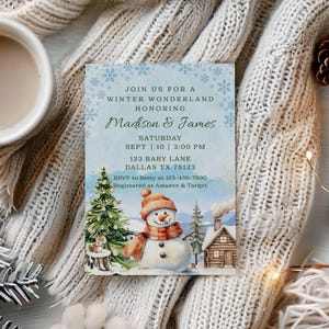 May include: A winter wonderland invitation with a watercolor snowman, pine tree, and cabin scene. The invitation reads "Join us for a Winter Wonderland honoring Madison & James, Saturday, Sept 10, 3:00 PM, 123 Baby Lane, Dallas TX 75123". The scene is set on a cream-colored knitted blanket, with a cup of coffee, pine cones, and a candle.