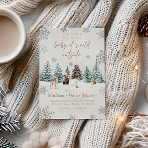May include: A baby shower invitation with the text "baby it's cold outside" and a winter forest scene. The invitation is surrounded by a cozy blanket, a cup of coffee, pine cones, and a lit candle. The invitation is for Madison and James Johnson.