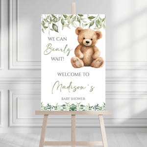 May include: A white welcome sign with green leafy branches and a brown teddy bear. The sign reads "We can Bearly wait! Welcome to Madison's Baby Shower".