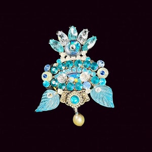 Può includere: Un ciondolo blu e bianco decorato con gioielli con una perla pendente. Il ciondolo presenta un motivo a occhio centrale circondato da gioielli blu e bianchi più piccoli.