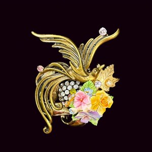 May include: A gold-tone brooch featuring a stylized bird with a floral design. The bird is adorned with rhinestones and the flowers are made of porcelain.