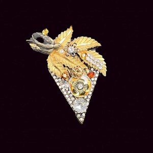 Könnte beinhalten: Eine goldfarbene Brosche in Dreiecksform mit einem Cluster aus Blättern, Blumen und Strasssteinen. Die Brosche hat eine schwarze und goldene Oberfläche.