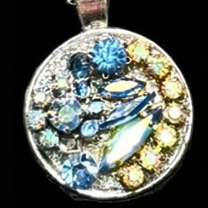 May include: A silver pendant with a circular design, featuring an assortment of colourful gemstones. The gemstones include shades of blue, yellow, and clear crystals, creating a sparkling effect. The pendant is suspended from a silver chain.