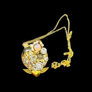 Può includere: Una collana d'oro con un ciondolo a forma di cuore adornato con cristalli scintillanti e una pietra rosa. Il ciondolo presenta delicate ali e un ciondolo a forma di cuore.
