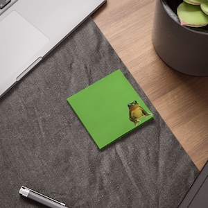 Op de afbeelding: Een groene sticky note met een kikker sticker erop. De kikker is groen en bruin met een witte buik. De sticky note ligt op een grijs oppervlak.