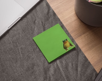 May include: A green sticky note with a frog sticker on it. The frog is green and brown with a white belly. The sticky note is on a grey surface.