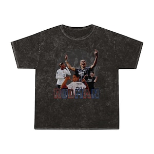 May include: A black t-shirt with a faded, distressed look. The shirt features a graphic of a group of football players celebrating a goal. The text "RODDMAN" is printed in red and blue letters across the bottom of the graphic.