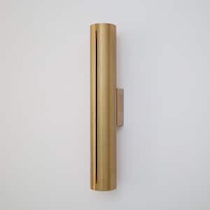 Puede incluir: Aplique de pared cilíndrico dorado con placa de montaje rectangular. El aplique tiene una ranura vertical en la parte delantera. La luminaria está montada en una pared blanca.