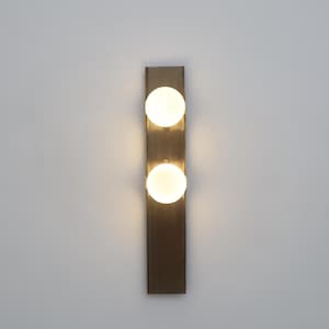 May include: A contemporary wall light with two spherical, frosted glass bulbs. The bulbs are mounted vertically on a rectangular, bronze-toned metal backplate. Suitable for interior lighting applications.