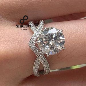 May include: A silver engagement ring with a large, round, clear gemstone. The band features a unique, intertwined design with smaller, sparkling stones. The ring is from SHRESVI JEWELS.
