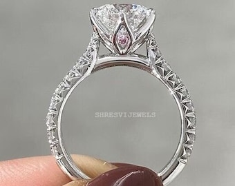 Peut inclure: Une bague de fiançailles en argent avec une grosse pierre centrale ronde et claire et un halo de pierres plus petites. La bande est également sertie de pierres plus petites et présente un design délicat et complexe. La bague est tenue par une main avec un vernis à ongles rouge.
