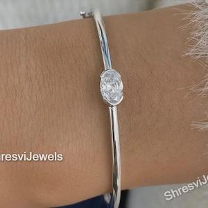 Puede incluir: Una pulsera de plata con una sola gema de corte ovalado. La pulsera tiene un diseño sencillo y elegante, ideal para el uso diario. La gema está engastada en un bisel y la pulsera tiene un acabado liso y pulido. El texto "ShresviJewels" es visible.