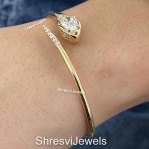 May include: A gold-toned cuff bracelet with a snake head design. The snake's head features a pear-shaped, clear gemstone. The bracelet's body is a smooth, curved band. The text "ShresviJewels" is visible.