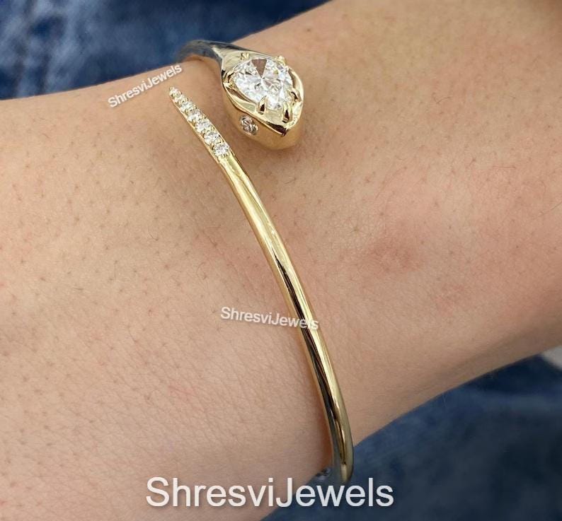 May include: A gold-toned cuff bracelet with a snake head design. The snake's head features a pear-shaped, clear gemstone. The bracelet's body is a smooth, curved band. The text "ShresviJewels" is visible.