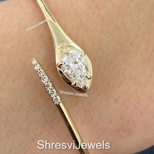May include: A gold-colored cuff bracelet with a pear-shaped diamond at one end and a line of smaller diamonds on the other. The bracelet is open and wraps around the wrist. The text "ShresviJewels" is visible.