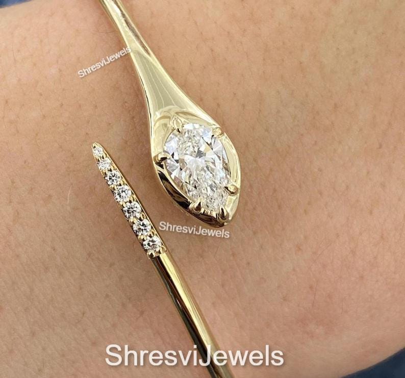 May include: A gold-colored cuff bracelet with a pear-shaped diamond at one end and a line of smaller diamonds on the other. The bracelet is open and wraps around the wrist. The text "ShresviJewels" is visible.