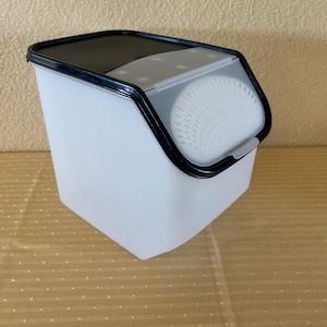 May include: A white plastic container with a black rim and lid. The container has a gray, perforated section on the front. The lid is black and the container is angled. The container is designed for kitchen use.