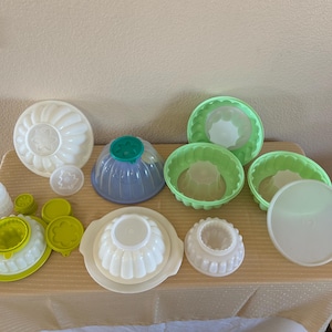 May include: A collection of various plastic molds in white, green, and blue. The molds are in different shapes and sizes, including round, fluted, and dome-shaped designs. Some have lids or decorative tops. Ideal for making desserts.