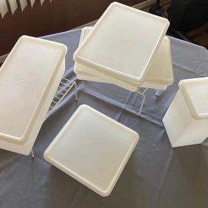 May include: A collection of white plastic food storage containers with lids. Some containers are rectangular, and others are square. The containers are on a gray surface.