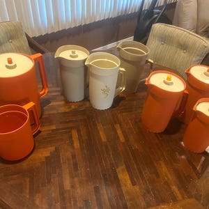 May include: A collection of vintage pitchers and a cup in various colors. The pitchers are orange, beige, and white, with lids and handles. Some have floral designs. The items are arranged on a dark wooden table.