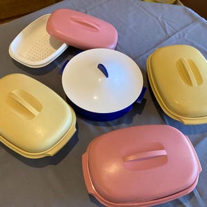 May include: A selection of vintage plastic food storage containers in various colours. The containers include pink, yellow, white, and blue lids and bases. Some have handles, and one has a perforated insert.