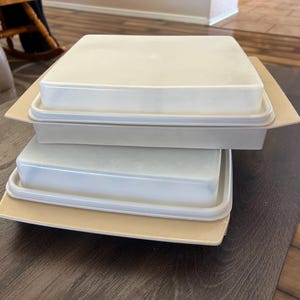 May include: Three stacked, off-white plastic food storage containers with lids. Each container sits on a beige, rectangular tray. The containers are on a dark brown table, with a glimpse of a wooden floor and a chair in the background.