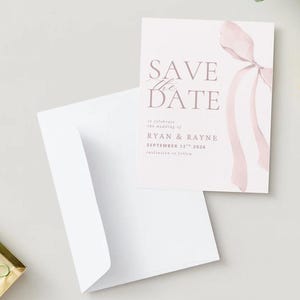 May include: A wedding Save the Date card with a pink watercolour ribbon design. The card displays the text "SAVE the DATE" and wedding details. A white envelope is open beside the card, with a gold tray of green paperclips in the corner.