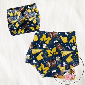 May include: A navy blue and yellow football-themed baby outfit set. The set includes a headband and shorts, both featuring a repeating pattern of footballs, the letter "M", foam fingers, and football helmets. The word "FOOTBALL" is also visible.
