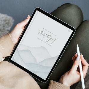 May include: A digital tablet displaying a journal cover with a watercolor background of blue and gray mountains. The cover text reads "Becoming That Girl: The Shadow Work Journal".