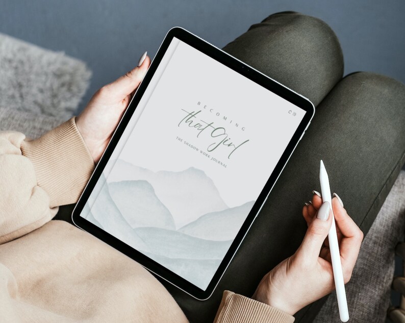 May include: A digital tablet displaying a journal cover with a watercolor background of blue and gray mountains. The cover text reads "Becoming That Girl: The Shadow Work Journal".