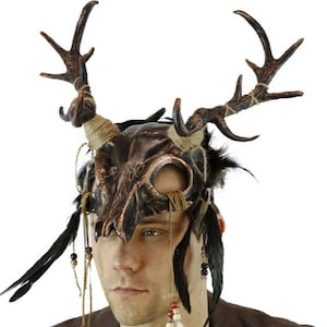 May include: A brown and black skull mask with antlers and feathers. The mask has a circular opening for the eyes and a long, pointed nose.