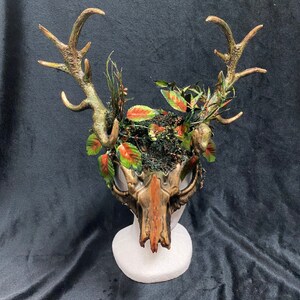 May include: A gold and brown deer skull mask with antlers and green and red foliage accents.