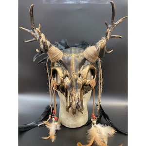 May include: A gold and brown deer skull mask with black feathers and brown leather straps. The mask has a distressed look and is decorated with beads.