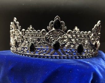 May include: A silver-toned crown with intricate filigree detailing and teardrop-shaped black gemstones. The crown sits on a blue velvet surface, showcasing its ornate design. This regal headpiece is suitable for special occasions.