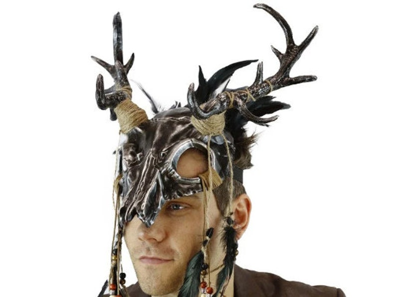 May include: A silver and brown skull mask with antlers and feathers. The mask has a tribal design and is worn by a person.
