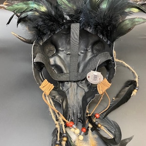 May include: Black skull mask with feathers, beads, and leather cords. The mask has a black elastic band for wearing.