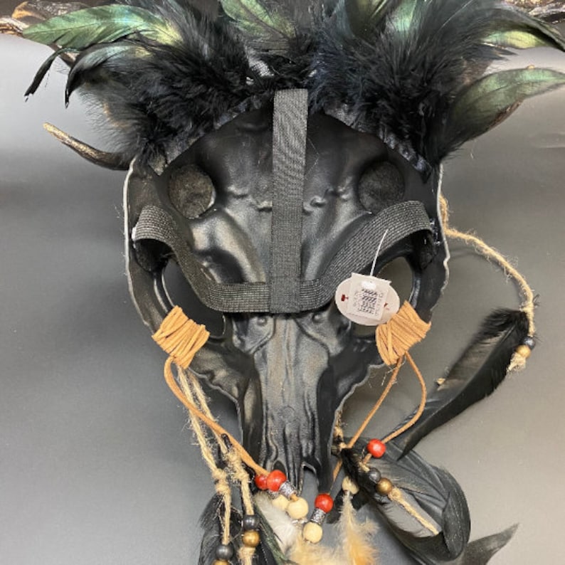 May include: Black skull mask with feathers, beads, and leather cords. The mask has a black elastic band for wearing.