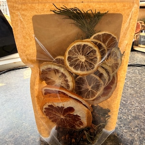 May include: A clear plastic bag filled with dried orange and lemon slices, cloves, and sprigs of rosemary. The bag has a brown paper backing and a clear window. The contents are visible through the window, showcasing the natural colors and textures of the ingredients.