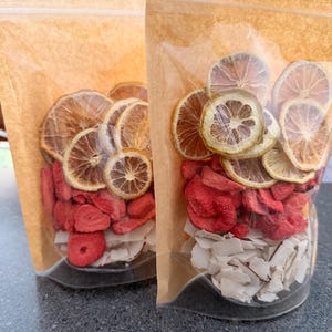 May include: Two clear plastic bags filled with dried fruit. The bags are sealed with a zipper closure and have a brown paper backing. The bags contain dried lemon slices, red strawberry pieces, and white coconut flakes. The bags are sitting on a dark gray surface.
