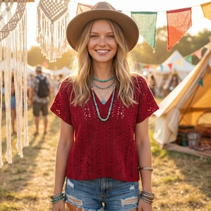 Puede incluir: Una mujer sonr&iacute;e, luciendo un top de ganchillo burdeos, pantalones cortos de mezclilla y un sombrero marr&oacute;n. Lleva collares y pulseras turquesas. El fondo muestra un festival con tiendas de campa&ntilde;a y banderines decorativos.