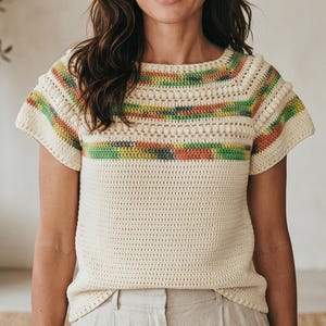May include: Cream-colored crochet top with short sleeves. The top features a horizontal striped pattern in shades of green, yellow, orange, and brown across the neckline and sleeves. The body of the top has a simple, open stitch design.