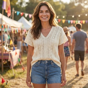 Puede incluir: Una mujer con un top de crochet crema con cuello en V y pantalones cortos de mezclilla sonr&iacute;e a la c&aacute;mara. El fondo muestra un festival con tiendas de campa&ntilde;a, banderas de colores y gente caminando.