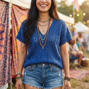 Puede incluir: Una persona lleva un top azul de ganchillo y pantalones cortos de mezclilla. Lleva varios collares y pulseras. El fondo muestra un ambiente de festival con tiendas de campa&ntilde;a y gente.