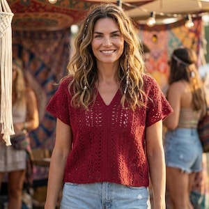 Puede incluir: Una mujer con un top de ganchillo burdeos con cuello en V y mangas cortas, combinado con shorts vaqueros azul claro. El fondo muestra un mercado de estilo bohemio con textiles coloridos y luces decorativas, lo que sugiere un festival o evento al aire libre.