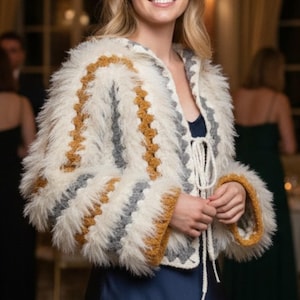 May include: A fluffy, cream-colored jacket with a crochet design in gray and mustard yellow. The jacket has long sleeves with mustard yellow cuffs and a tie closure at the neckline. The jacket is worn over a navy blue top.