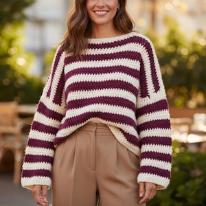 May include: A cream and burgundy striped crochet sweater. The sweater has a wide, boat neck and long sleeves. The stripes are horizontal and evenly spaced. The model is wearing tan trousers.